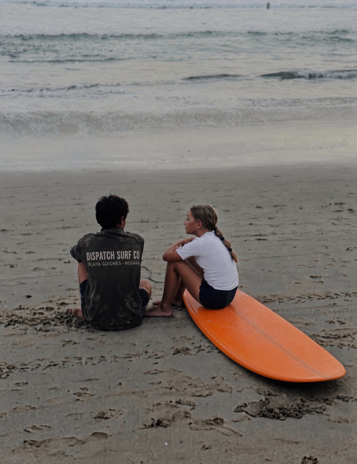 Two kids with an orange longboard at the water's edge at Playa Guiones